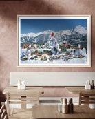A large framed art print depicting a winter mountain landscape with a snowy outdoor dining scene, displayed on a textured brown wall above a wooden dining table and chairs.