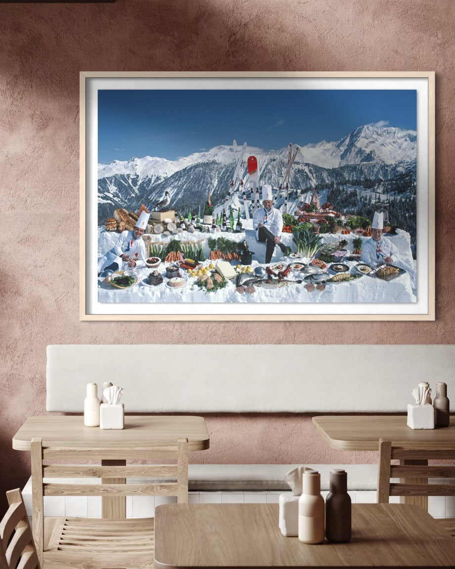 A large framed art print depicting a winter mountain landscape with a snowy outdoor dining scene, displayed on a textured brown wall above a wooden dining table and chairs.