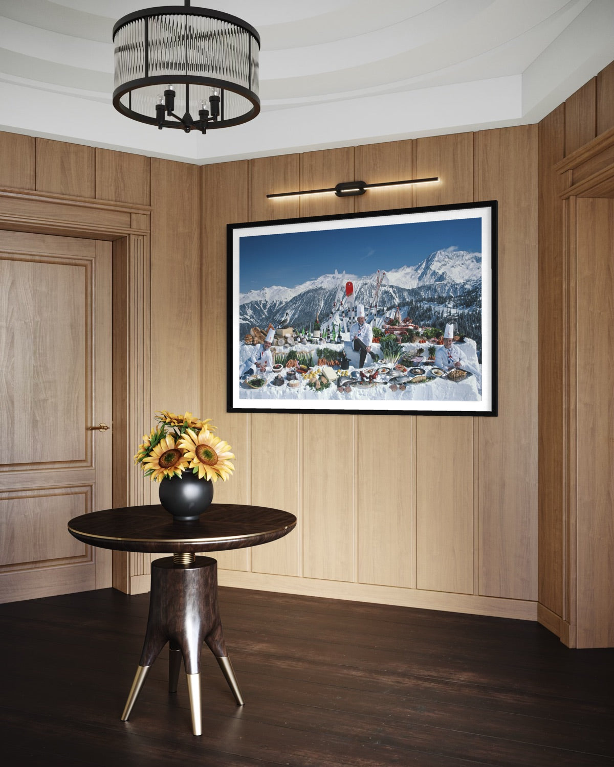 A framed art print depicting a snowy mountain village under a blue sky, displayed on a wood-paneled wall in a modern interior setting with a dark wood table and sunflowers in the foreground.
