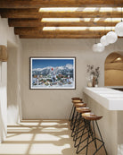 A framed landscape art print depicting a snowy mountain village under a blue sky, displayed on a beige wall in a modern rustic kitchen setting with wooden ceiling beams, pendant lights, and bar stools.