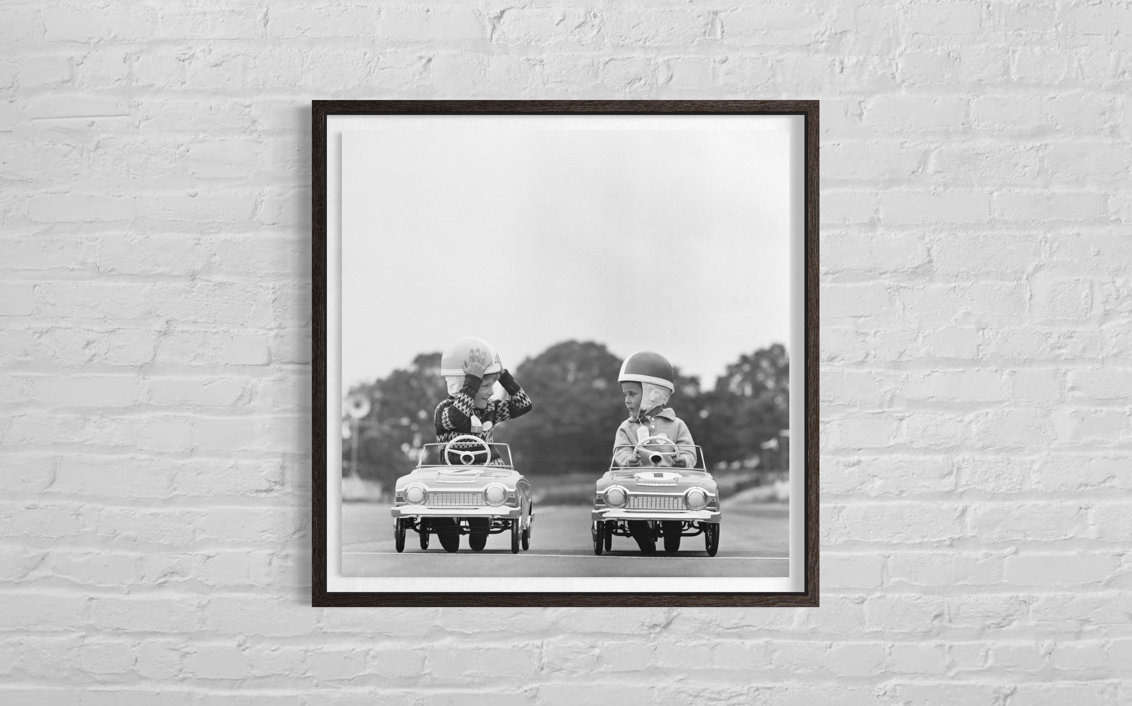 Framed black and white photograph of two children in toy cars on a brick wall.