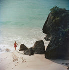 A vintage-style photograph of a beach scene with a person in a red swimsuit walking near the shoreline, large dark rocks, pale sand, and turquoise ocean water. The Picturalist Wall Art and Photography for Designed Interiors. Slim Aarons Collection, Coastal. Getty Images Gallery. 