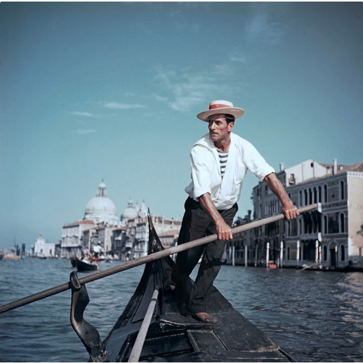 A man rowing a gondola on a canal in Venice, with historic buildings and a domed cathedral visible in the background under a blue sky. The Picturalist Wall Art and Photography for Designed Interiors. Slim Aarons Collection, Summer Travel. Getty Images Gallery. 