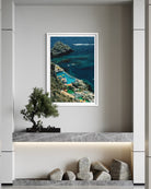 A framed vertical art print depicting a scenic coastal cove with turquoise blue water, rocky cliffs, and green hills, displayed on a light gray wall above a marble console with a small tree and stones.