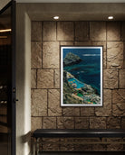 A framed vertical art print depicting a scenic coastal cove with turquoise water, rocky cliffs, and green vegetation, displayed on a textured stone wall above a dark bench in an interior setting.
