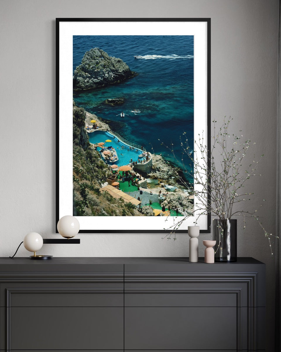 A framed vertical art print of a coastal resort with turquoise pools and rocky cliffs, displayed on a dark gray console table against a light gray wall with decorative vases.