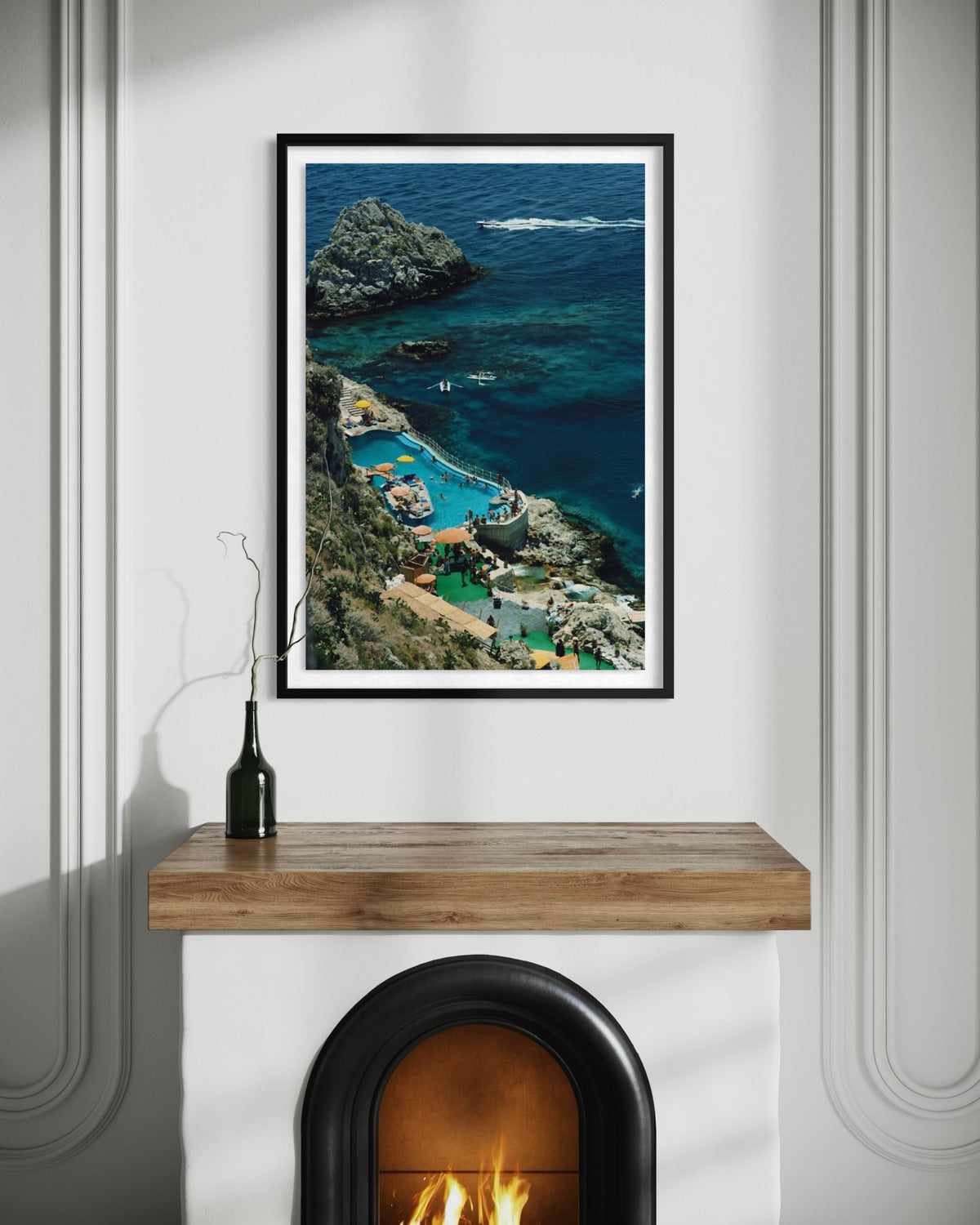 A framed vertical art print depicting a scenic coastal cove with turquoise water, rocky cliffs, and a villa, displayed on a white wall above a wooden mantel with a fireplace visible below.