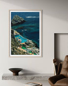 A framed vertical art print depicting a coastal cove with deep blue ocean, rocky cliffs, and turquoise pools, displayed on a light gray wall above a mantel in a modern room setting.