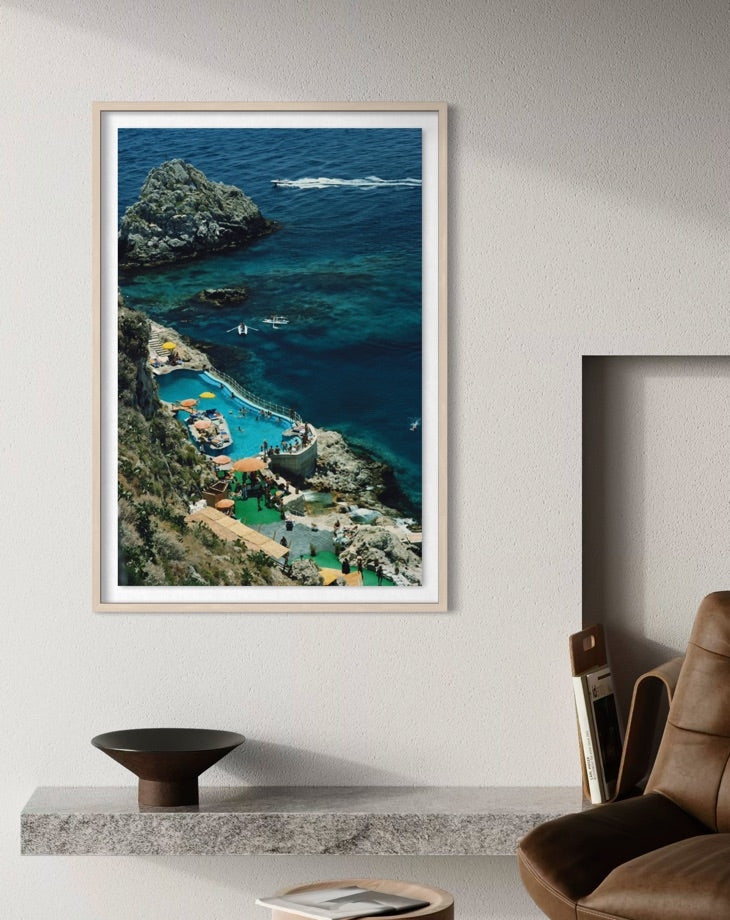 A framed vertical art print depicting a coastal cove with deep blue ocean, rocky cliffs, and turquoise pools, displayed on a light gray wall above a mantel in a modern room setting.