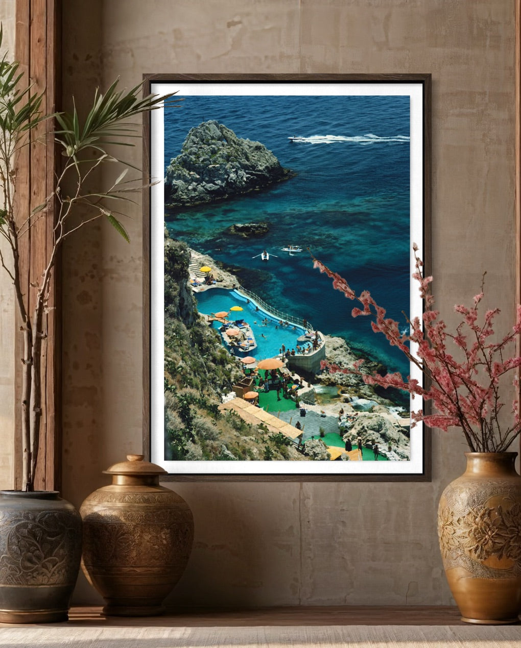 A framed vertical art print depicting a scenic coastal landscape with turquoise and deep blue ocean, rocky cliffs, and a resort area, displayed on a shelf against a textured wall with decorative vases.