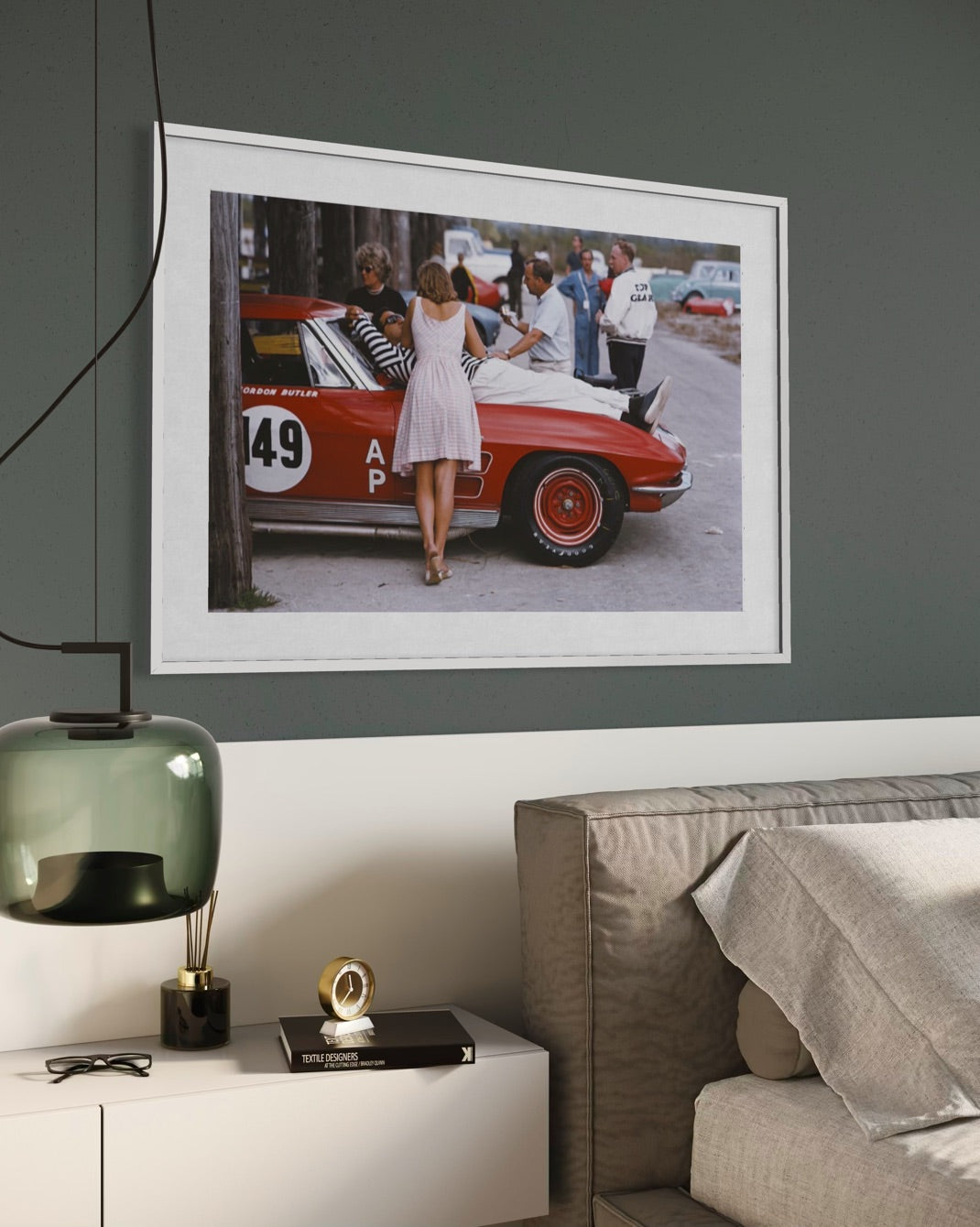 A framed art print depicting a vintage red race car and people on a street, displayed on a dark gray wall above a white sideboard with a green glass lamp and a beige sofa.