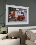 A framed art print depicting a vintage red race car and people on a street, displayed on a dark gray wall above a white sideboard with a green glass lamp and a beige sofa.