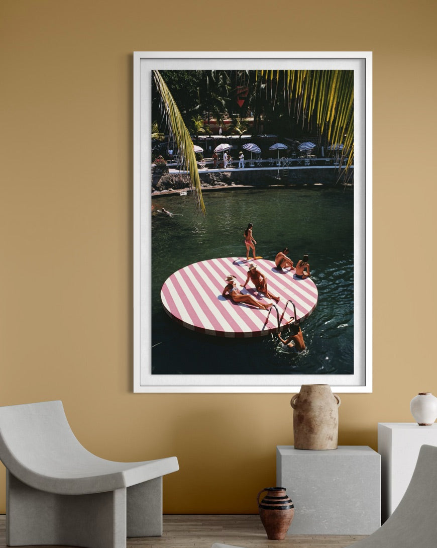 A framed vertical art print depicting a retro scene of people relaxing on a pink and white striped circular pool float in a tropical pool setting, displayed on a warm beige wall in a modern interior setting. The Picturalist Wall Art and Photography for Designed Interiors. Slim Aarons Collection, Costal. Getty Images Gallery. 