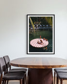 A framed vertical art print depicting a woman relaxing on a pink and white striped circular float in a tropical pool, displayed on a light gray wall above a wooden dining table and chairs. The Picturalist Wall Art and Photography for Designed Interiors. Slim Aarons Collection, Coastal. Getty Images Gallery. 