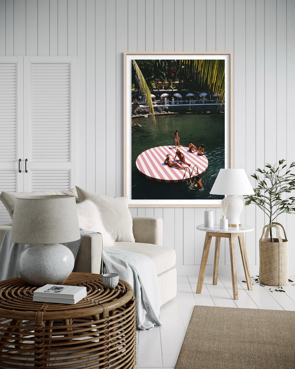 A framed vertical art print featuring a tropical pool scene with a pink-and-white striped float, displayed on a white paneled wall above a modern living room setup with a sofa, side table, lamp, and plant. The Picturalist Wall Art and Photography for Designed Interiors. Slim Aarons Collection, Coastal. Getty Images Gallery. 