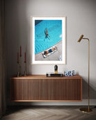 A framed art print depicting an aerial view of a person swimming in a blue pool next to another person sunbathing, styled on a gray wall above a wooden console table in a modern living room setting. The Picturalist Slim Aarons Collection.