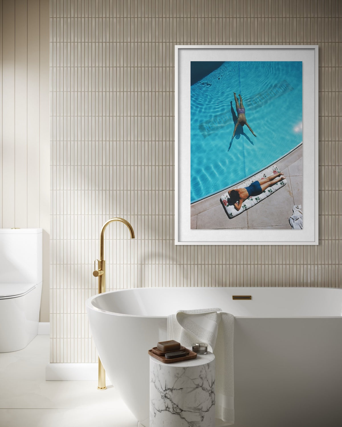 A framed art print depicting a person floating in a swimming pool next to a person sunbathing on a pool deck, displayed on a beige tiled wall above a modern white bathtub with a gold faucet.  The Picturalist Slim Aarons Collection