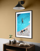 A framed art print depicting a person swimming in a bright blue pool, styled on a wooden console table against a warm beige wall under a black wall sconce.  The Picturalist Slim Aarons Collection