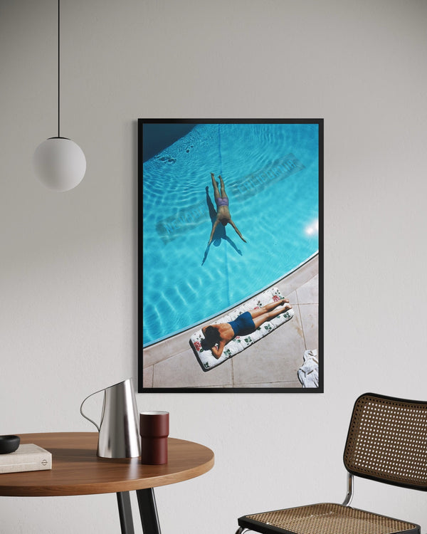 A framed vertical art print depicting a high-angle view of a person swimming in a bright blue pool next to a person lying on a lounge chair, displayed on a light gray wall in a modern room setting.  The Picturalist Slim Aarons Collection