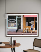A framed art print depicting a Parisian street scene with a coffee shop and outdoor seating, displayed on a light gray wall above a wooden table with a lamp and chair. The Picturalist Slim Aarons Collection. 