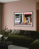 A framed art print depicting a Parisian street scene with a cafe and a person, displayed on a dusty pink wall above a dark green velvet sofa in a modern living room setting. The Picturalist Slim Aarons Collection. 