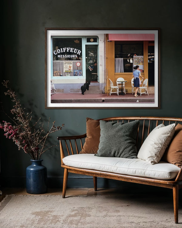 A framed art print depicting a Parisian street scene with a coffee shop and outdoor seating, displayed on a dark green wall above a wooden bench with cushions. The Picturalist Slim Aarons Collection. 