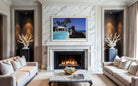 A framed art print depicting a tropical beach scene with palm trees, a pool, and a black statue, displayed on a white marble wall above a fireplace in a modern living room with beige sofas. The Picturalist Slim Aarons Collection. 