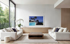 A framed art print depicting a tropical beach scene with a pool and statue, displayed on a light wood console in a modern, bright living room with white sofas and a wooden coffee table. The Picturalist Slim Aarons Collection. 