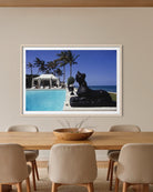 A framed art print depicting a tropical resort scene with a swimming pool, palm trees, a black lion statue, and the ocean, displayed on a beige wall above a wooden dining table with chairs. The Picturalist Slim Aarons Collection. 