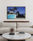 A framed art print depicting a luxurious tropical beach scene with a swimming pool, palm trees, and a black statue, displayed on a white wall above a modern beige sofa with a marble-top coffee table. The Picturalist Slim Aarons Collection. 