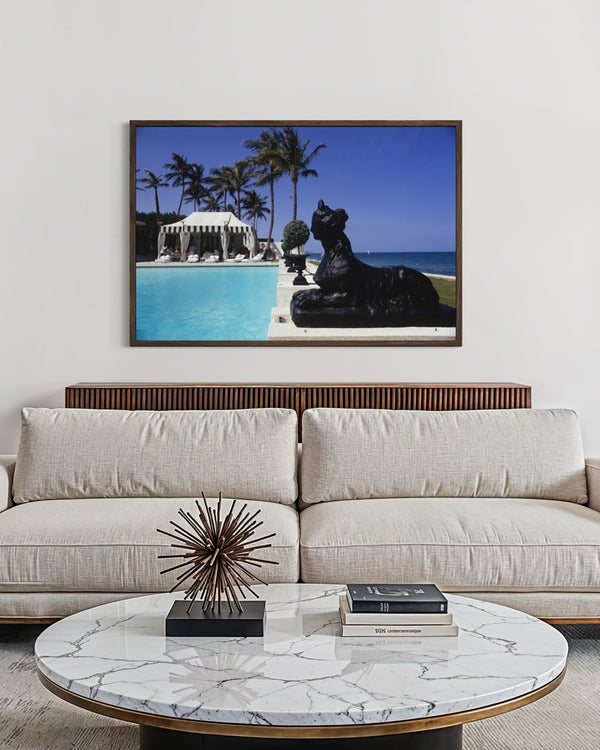 A framed art print depicting a luxurious tropical beach scene with a swimming pool, palm trees, and a black statue, displayed on a white wall above a modern beige sofa with a marble-top coffee table. The Picturalist Slim Aarons Collection. 