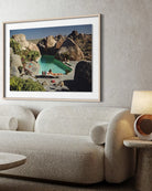 A framed art print depicting a scenic desert pool surrounded by large rocks, displayed on a textured light gray wall above a modern beige boucle sofa with a lamp on a side table. The Picturalist Slim Aarons Collection. 