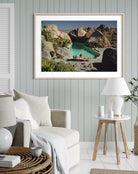 A framed art print depicting a scenic desert poolside landscape with large boulders and a turquoise pool, styled in a modern living room setting above a beige sofa.