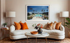 A framed art print depicting a tropical resort pool scene hangs on a beige wall above a white curved sofa with orange and patterned pillows in a modern living room setting. The Picturalist Slim Aarons Collection. 