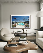A framed art print depicting a luxurious tropical poolside scene with palm trees and a blue sky, displayed on a light gray wall in a modern, elegant living room setting. The Picturalist Slim Aarons Collection. 