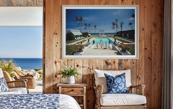 A framed art print depicting a tropical resort pool scene with palm trees and a blue sky, displayed on a rustic wooden wall above a wicker armchair in a styled bedroom setting. The Picturalist Slim Aarons Collection. 