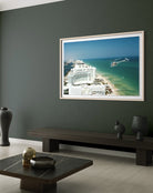 A framed photographic print of a coastal city and ocean scene hangs on a dark green wall above a long bench in a modern living room setting. The Picturalist Slim Aarons Collection. 