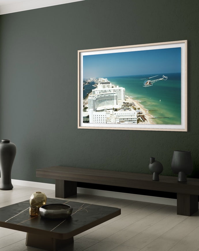 A framed photographic print of a coastal city and ocean scene hangs on a dark green wall above a long bench in a modern living room setting. The Picturalist Slim Aarons Collection. 