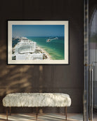 A framed coastal photograph hanging on a dark wood-paneled wall above a white faux-fur bench. The print shows a beach, turquoise ocean, and white buildings, with a black frame and white mat. The Picturalist Slim Aarons Collection. 