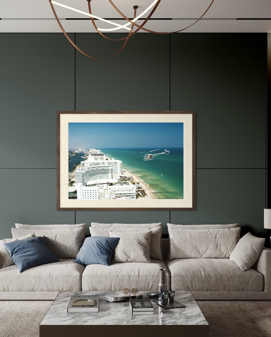 A framed art print depicting an aerial view of a tropical beach and cruise ships, displayed on a dark paneled wall above a modern light gray sofa in a living room setting. The Picturalist Slim Aarons Collection. 