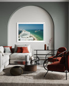 A framed art print depicting a coastal cruise ship scene in turquoise water, displayed on a gray wall in a modern living room setting with a white sofa, red accent pillows, and a red armchair. The Picturalist Slim Aarons Collection. 