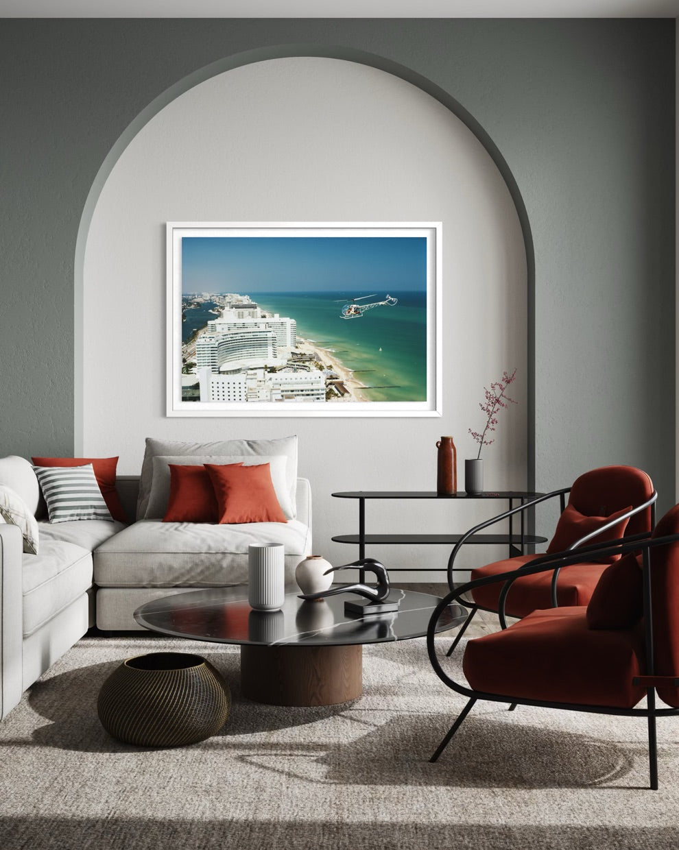 A framed art print depicting a coastal cruise ship scene in turquoise water, displayed on a gray wall in a modern living room setting with a white sofa, red accent pillows, and a red armchair. The Picturalist Slim Aarons Collection. 