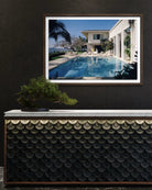 A framed art print depicting a luxurious outdoor poolside scene with a modern house, palm trees, and blue sky, displayed on a dark wall above a decorative console table with a bonsai tree.  The Picturalist Wall Art and Photography. Slim Aarons Collection. 
