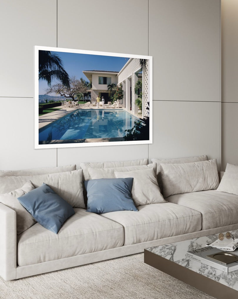 A framed art print depicting a luxurious outdoor villa with a swimming pool, displayed on a light gray paneled wall above a modern beige sofa with blue pillows.  The Picturalist Wall Art and Photography. Slim Aarons Collection. 