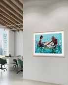 A framed art print depicting a couple sitting at a table by a turquoise ocean, displayed on a white wall in a modern office interior with wooden ceiling beams and office chairs. The Picturalist Wall Art and Photography. Slim Aarons Collection. Poolside. 
