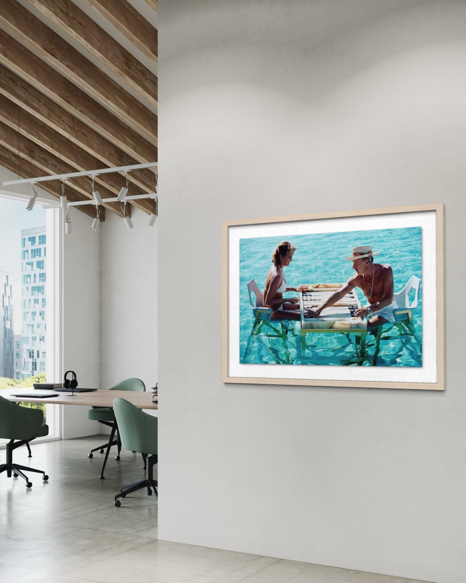 A framed art print depicting a couple sitting at a table by a turquoise ocean, displayed on a white wall in a modern office interior with wooden ceiling beams and office chairs. The Picturalist Wall Art and Photography. Slim Aarons Collection. Poolside. 