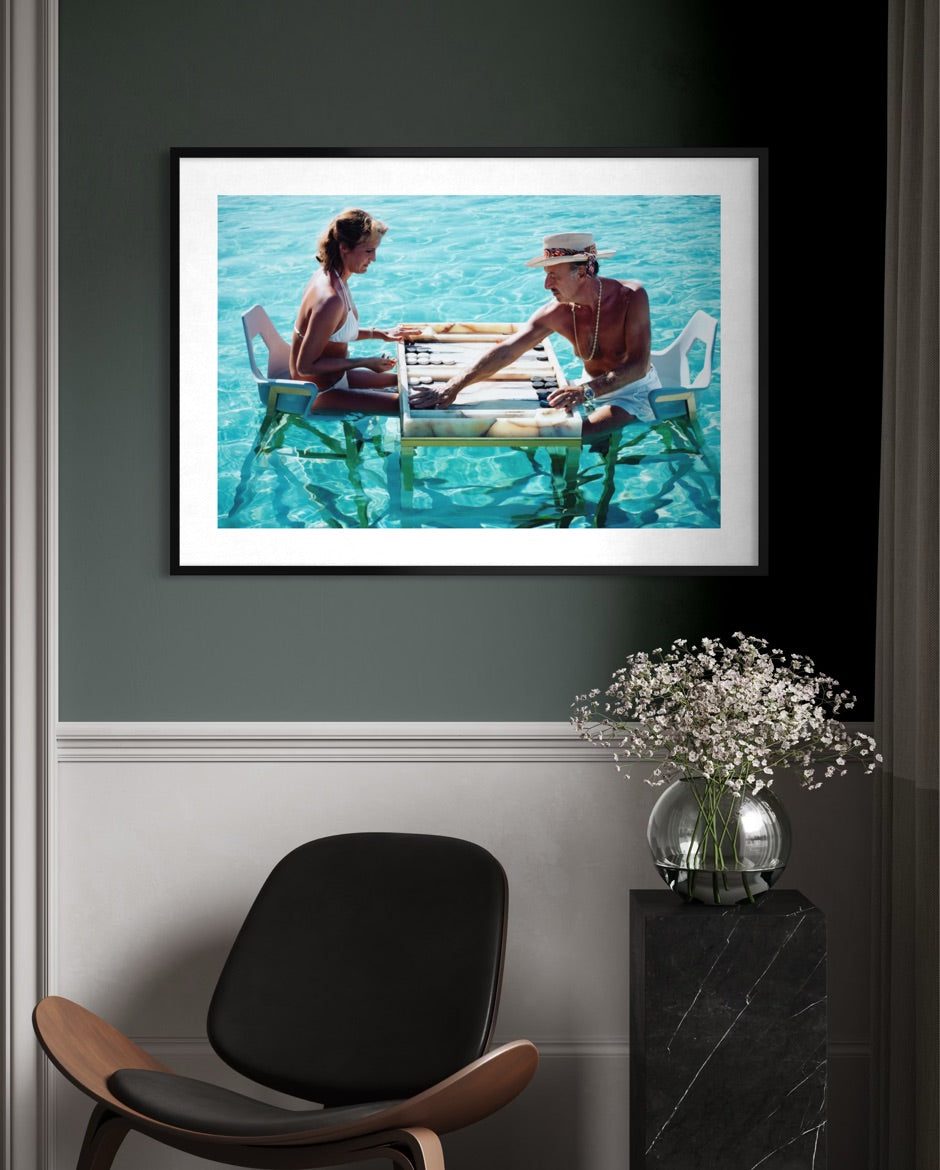A framed art print depicting a couple sitting on a wooden dock in turquoise water, displayed on a dark gray wall above a modern black and wood chair with a vase of baby's breath on a marble side table. The Picturalist Wall Art and Photography. Slim Aarons Collection. Poolside. 