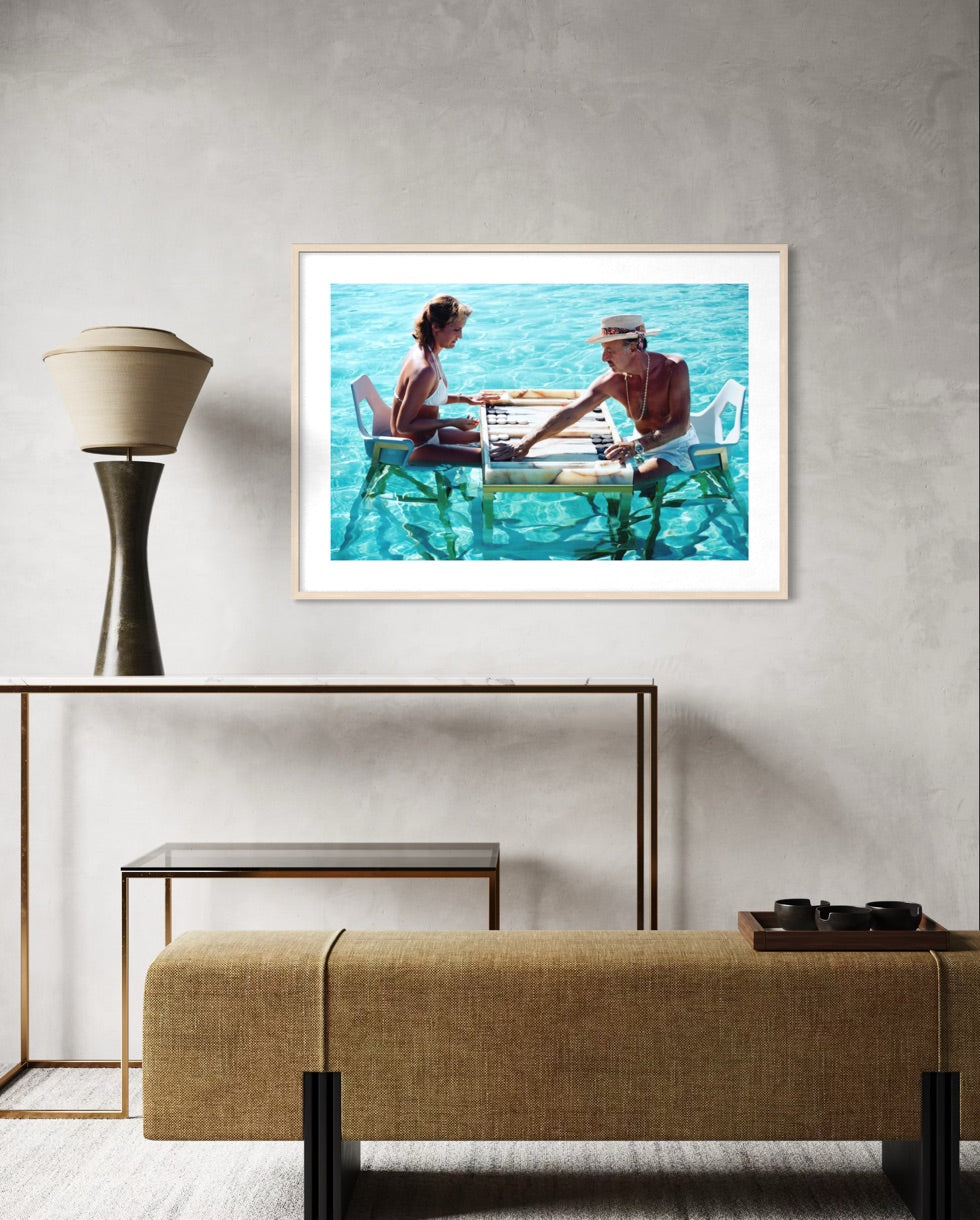 A framed art print depicting a couple sitting at a table over clear turquoise water, displayed on a textured light gray wall above a modern console table and textured bench. The Picturalist Wall Art and Photography. Slim Aarons Collection. Poolside. 