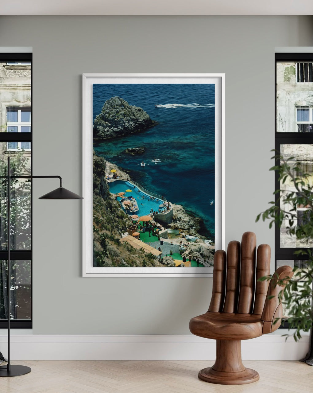 A framed vertical art print of a coastal resort with turquoise pools and rocky cliffs, displayed on a light gray wall in a modern room with a wooden hand-shaped chair and windows.