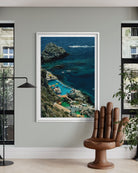A framed vertical art print of a coastal resort with turquoise pools and rocky cliffs, displayed on a light gray wall in a modern room with a wooden hand-shaped chair and windows.
